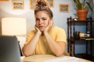 Upset overworked female sitting at table with laptop working remotely, feeling tired trying to catch deadline while finishing project, resting her chin on hands looking at camera with frustration