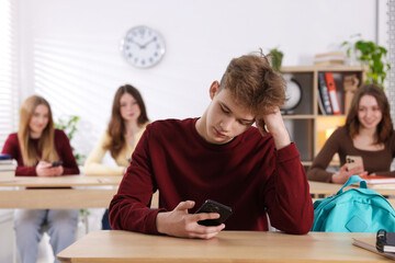 Cyber bullying. Upset teenage boy with smartphone at desk in classroom