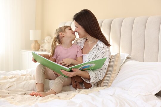 Bedtime. Happy little daughter reading book with her mother on bed at home