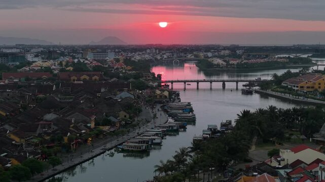 Drone view of Hoi An, Vietnam in the early morning showing the beautiful sunrise. Boats float on the Thu Bon River beside yellow historic buildings . Peaceful and scenic.