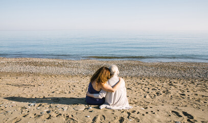 Rear view of an elderly mother and her adult daughter sitting close together on a sandy beach, wrapped in a warm embrace. The peaceful sea and shared moment evoke themes of connection, love, and gener