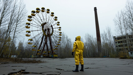 A person in a yellow hazmat suit and gas mask stands in front of an abandoned Ferris wheel, surrounded by leafless trees and signs of decay in a desolate, overcast environment	