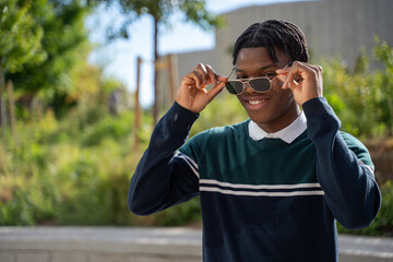 Portrait of a smiling young student adjusting his sunglasses while standing outdoors in a park
