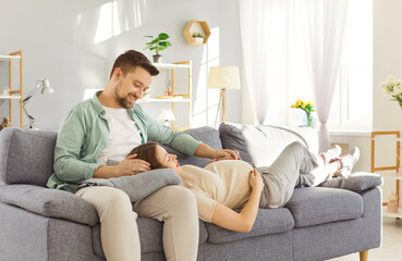 Obraz premium Young man sitting on sofa in living room at home talking with pregnant wife touching her big belly. Happy couple expecting a baby preparing to become parents. Pregnancy and parenthood concept.
