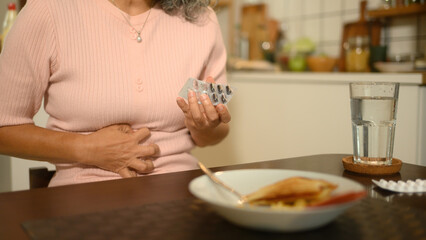Close up image of an elderly woman holding a digestive medicine and a glass of water on the table,...