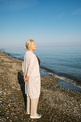 Elderly woman stands on a quiet beach with hands behind her back, looking at the sea. The calm...