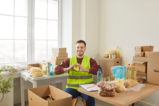 Cheerful smiling young man volunteer making heart gesture by hands volunteering in charitable foundation. Male person sitting in charity center. Humanitarian aid, altruism and social help concept.