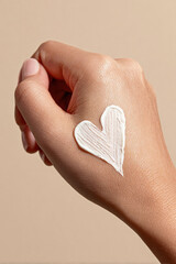 Close-up of hand with heart-shaped cream on skin, symbolizing skincare, softness, and self-care, against a neutral beige background with natural lighting.