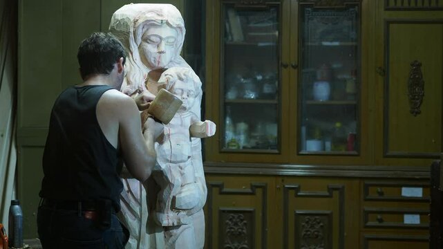Professional sculptor carefully chiseling intricate details of wooden religious statue using wood mallet anc chisel tools in controlled workshop environment, capturing precision, slow motion