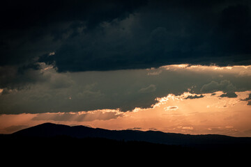 Dark storm clouds over mountain hills with soft light breaking through at sunset. A moody and atmospheric landscape capturing nature’s power, depth, and dramatic beauty