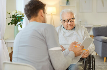 Male nurse sitting back to camera, holding tablet, showing results of analysis to handicapped senior man in wheelchair, explains prescription, takes care of him while being on rehabilitation