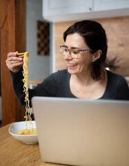 Happy Woman Eating Noodles And Working With A Laptop At The Kitchen Table