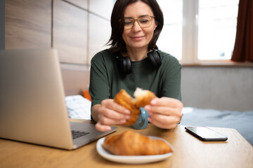 Pretty Woman Enjoys A Croissant For Breakfast While Checking Social Media On Laptop