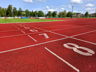 Bright red running track showing numbered lanes with crisp white numerals. The textured surface and clear markings lead toward verdant grass, distant trees and a vibrant blue sky above.