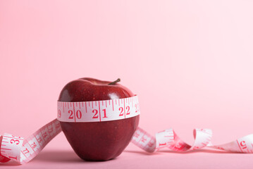 A red apple surrounded by a tape measure on a pink background. Weight control concept.