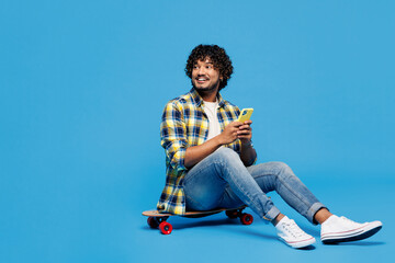 Full body young Indian man in yellow checkered shirt casual clothes sit on skateboard pennyboard...