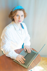 A beautiful young woman is working or playing or shopping on a laptop in a stylish white shirt and a hair band in the shape of the Russian national kokoshnik headdress