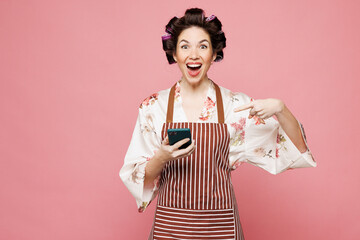 Young housewife housekeeper chef cook baker woman wears brown apron hold in hand use point finger on mobile cell phone isolated on pastel pink color background studio portrait. Cooking food concept.