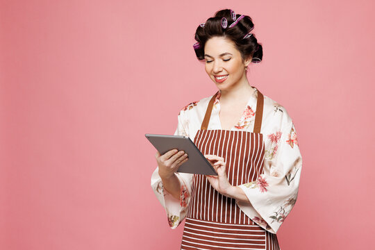 Young housewife housekeeper chef cook baker woman wear brown apron holding look for recipe on digital tablet pc computer isolated on pastel pink color background studio portrait. Cooking food concept.
