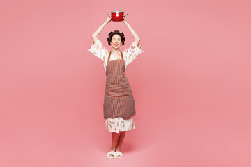 Full body young housewife housekeeper chef cook baker woman wear brown apron hold in hand red saucepan above head isolated on pastel light pink color background studio portrait. Cooking food concept.