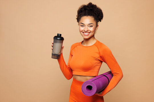 Young strong fitness trainer instructor sporty woman sportswoman in orange clothes spend time in home gym hold yoga mat protein shake isolated on plain beige background Workout sport fit abs concept