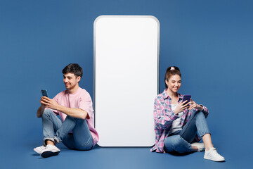 Plakat Full body young happy couple two friends family man woman wear pink casual clothes together sit near big huge blank screen mobile cell phone with area use smartphone isolated on plain blue background.