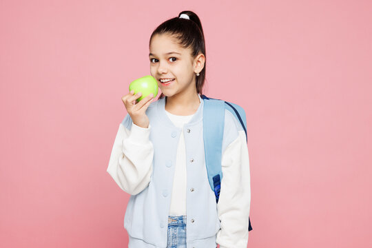 Young smiling happy school girl 8-9 years old wear blue casual clothes backpack hold eat bite apple look camera isolated on plain pink background children portrait. Childhood kids education concept.