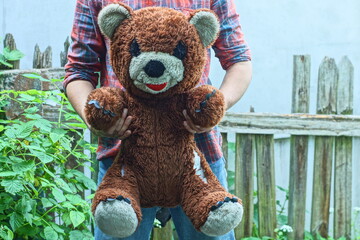 man in checkered shirt and jeans holding in hands one big old toy soft plush brown baby gift with black eyes and nose and white ears and face torn teddy bear outside