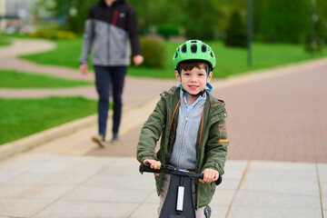 Obraz premium child riding scooter in park, wearing helmet outdoors. smiling boy enjoying leisure time on sunny day, embodying joy and safety. parent in background simulates supervision, relaxation