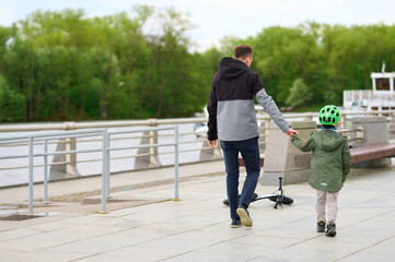 father and child holding hands walking riverside path exploring urban nature bonding over scooter...