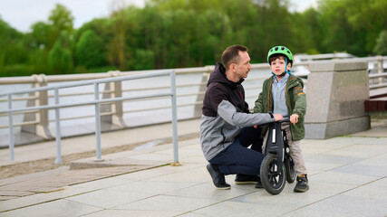 father teaching son to ride balance bike outdoors in park. man squatting beside young child wearing helmet. family bonding activity on clear day with nature backdrop