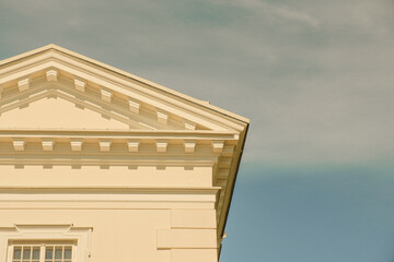 Close-up of a neoclassical triangular pediment and dentil cornice on a pale yellow facade. The architectural detail is bathed in warm sunlight.