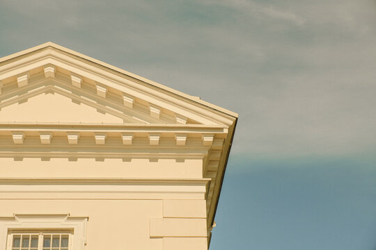 Close-up of a neoclassical triangular pediment and dentil cornice on a pale yellow facade. The architectural detail is bathed in warm sunlight.