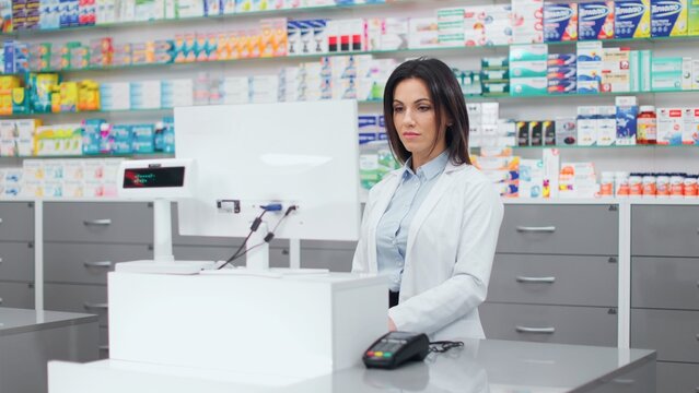 Female pharmacist looking at computer monitor. Woman pharmacist working with cash register. Woman in white robe standing at background of showcase with medicine. Caucasian pharmacist in drugstore.