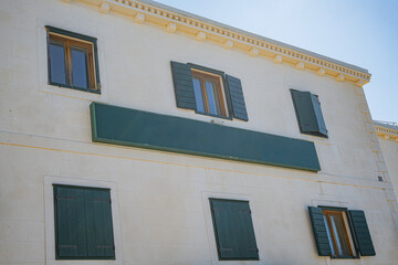 Facade of a traditional European stone building, featuring green wooden shutters on its multiple windows, all bathed in bright sunlight under a clear blue sky.