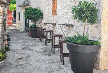 A cozy outdoor bar setup features wooden stools and small tables along a rustic stone wall, adorned with potted trees and greenery, evoking a charming Mediterranean ambiance.