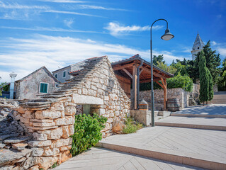 Sunlit street scene in Primosten, Croatia, featuring traditional stone architecture, a distinctive bell tower, and lush Mediterranean greenery under a dynamic blue sky with wispy clouds.