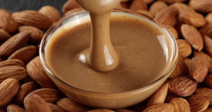 Almond nut butter is being scooped with a spoon. The almond butter drips from the spoon into a glass bowl on a background of almond nuts
