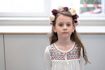 Cute girl in traditional Ukrainian embroidered shirt and flower wreath standing near large light...
