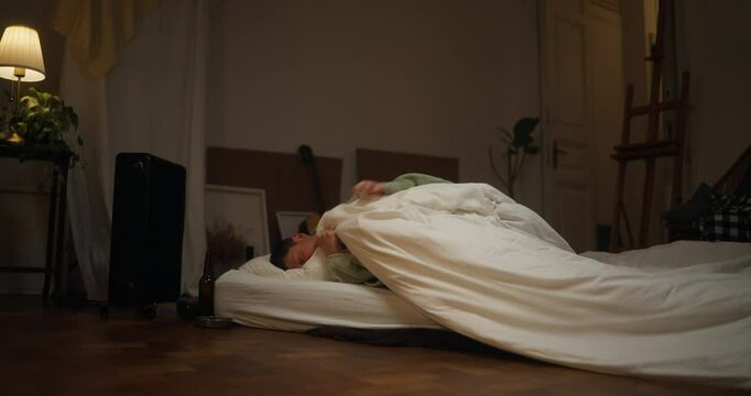 Tired male artist covers himself with a blanket and lies on a mattress near an ashtray at home. Trying to find peace and rest after a creative failure