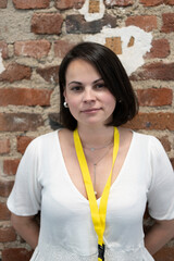 Beautiful dark-haired woman in a white blouse with a yellow lanyard badge, smiling confidently while standing against a brick wall. Business and professional concept