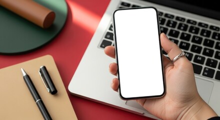 Hand holding a blank screen smartphone over a laptop with a notebook and pen on a red surface