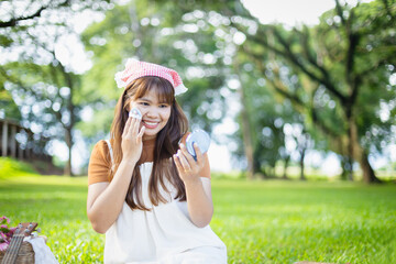 Young woman enjoys leisure time at picnic in park with green trees around as she smiles while applying makeup using compact mirror, capturing happy people concept