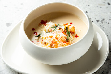 Creamy gourmet soup garnished with shredded cheese, herbs, and red sauce, served in a white bowl on a plate over a speckled surface