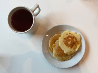 Sweet breakfast with pancakes topped with maple syrup and a cup of black tea, served in white tableware on a white table.