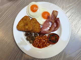 Traditional English breakfast on a white plate on a wooden table: 2 hash browns, 2 slices of bacon, baked beans, mushrooms, half a tomato and 2 fried eggs.