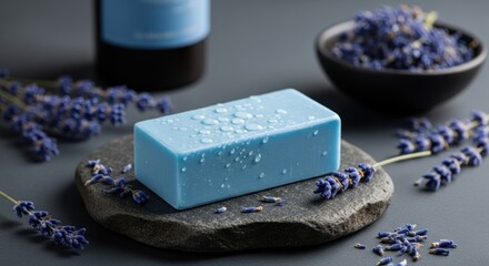 A light blue bar of soap with water droplets on a stone slab surrounded by lavender and a dark bottle