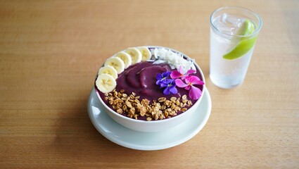 Acai smoothie bowl topped with banana, granola, coconut flakes, and edible flowers, served on a wooden table beside a glass of infused water.