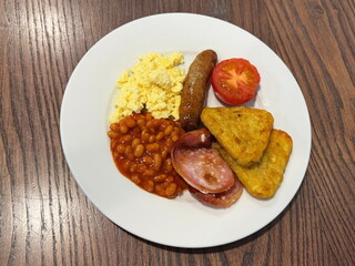 Traditional English breakfast on a white plate on a wooden table: 2 hash browns, 2 slices of bacon, baked beans, scrambled eggs, half a tomato, and a Lincolnshire sausage.
