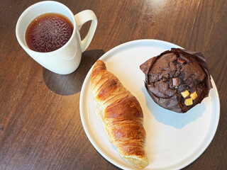 Black tea, croissant and chocolate muffin with chocolate chunks on white plate, sweet breakfast on dark wood table.